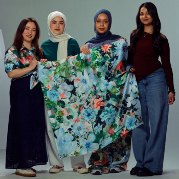 Four women stand together holding open a large floral-print garment, posed against a plain grey studio background.